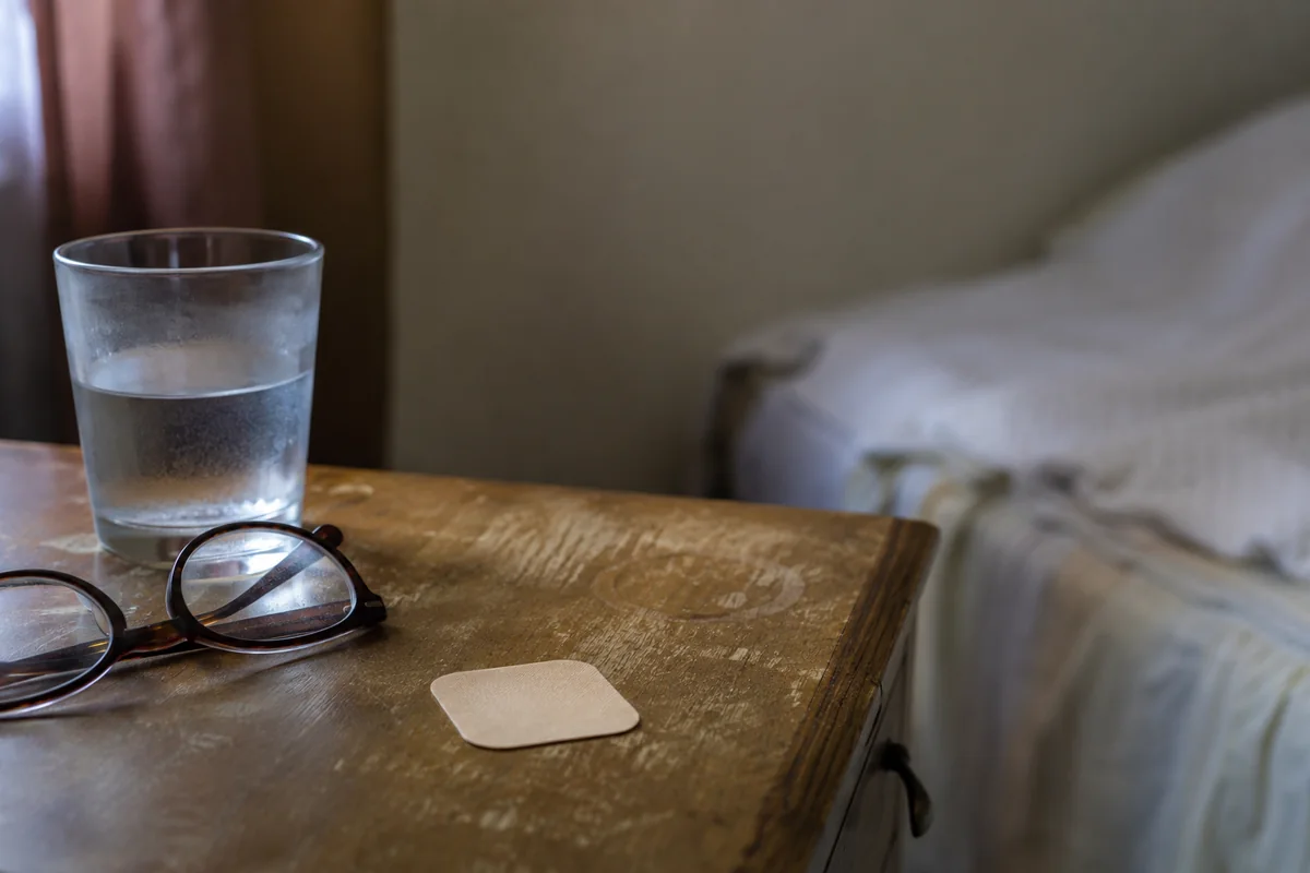 A small unbranded adhesive patch resting on a wooden nightstand beside a glass of water and reading glasses.
